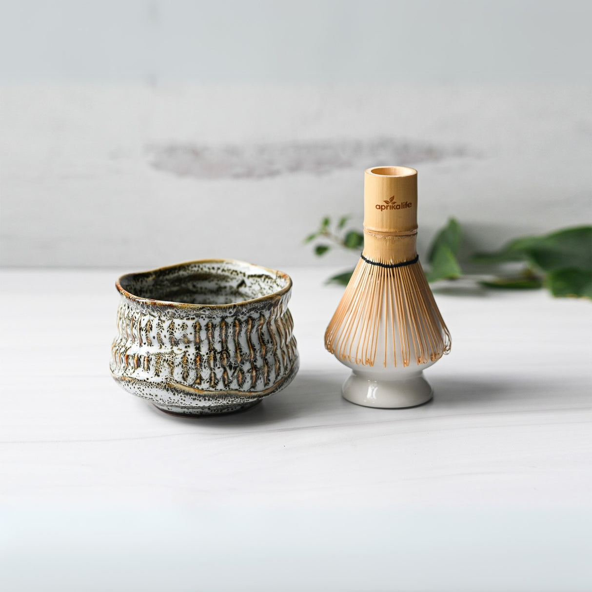 The White Ceramic Meisen Matcha Bowl and Bamboo Matcha Whisk from the Organic Japanese Matcha Set rest elegantly on a light surface, complemented by green foliage in the background.