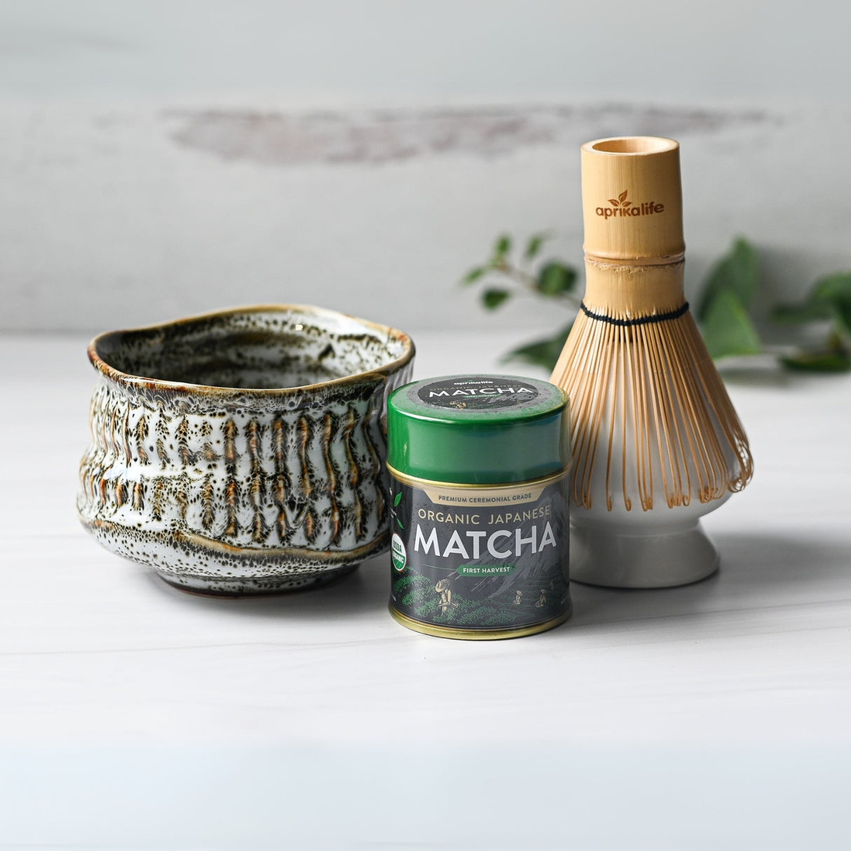 The White Ceramic Meisen Matcha Bowl, Bamboo Matcha Whisk, Whisk Holder, and Organic Japanese Matcha Set create the perfect matcha tea set on a light surface with a leafy background.