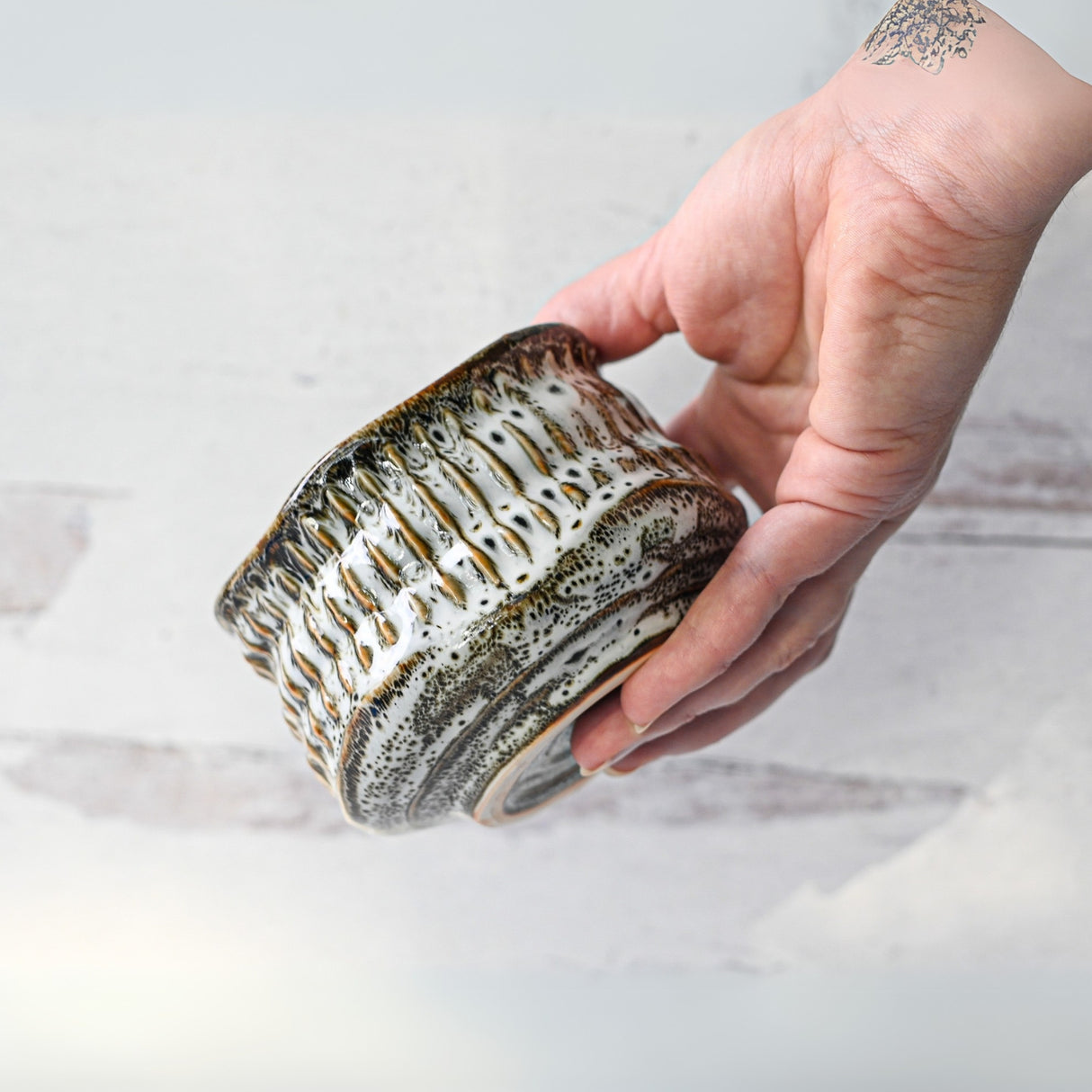 A hand holds the White Ceramic Meisen Matcha Bowl with a textured, speckled glaze against a light background, seamlessly complementing the Bamboo Matcha Whisk in any elegant matcha tea setting.