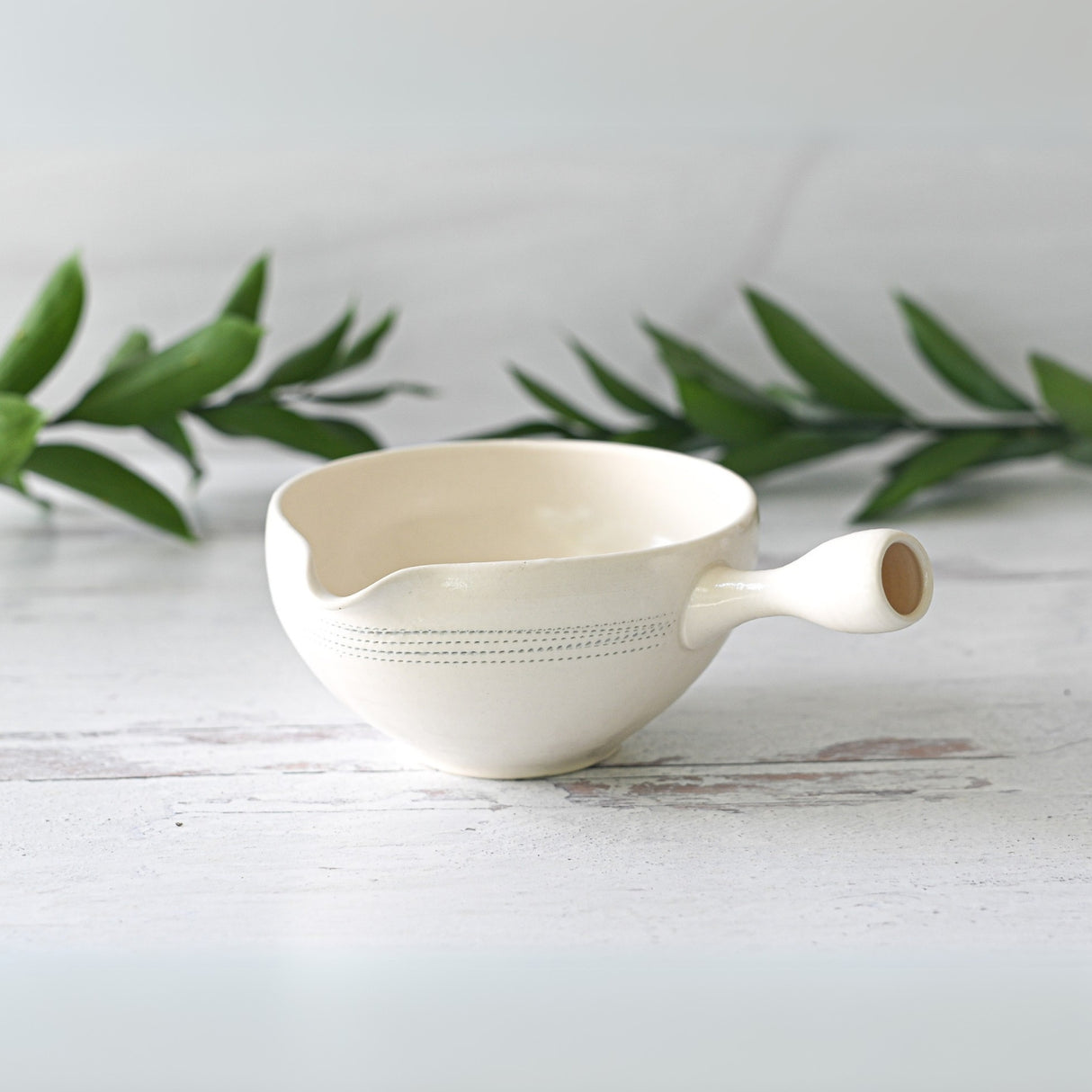 A White Striped Ceramic Matcha Bowl with Spout sits gracefully on a white surface, surrounded by green leaves, evoking a peaceful matcha tea ceremony vibe. It perfectly complements your Bamboo Matcha Whisk during serene tea preparation moments.