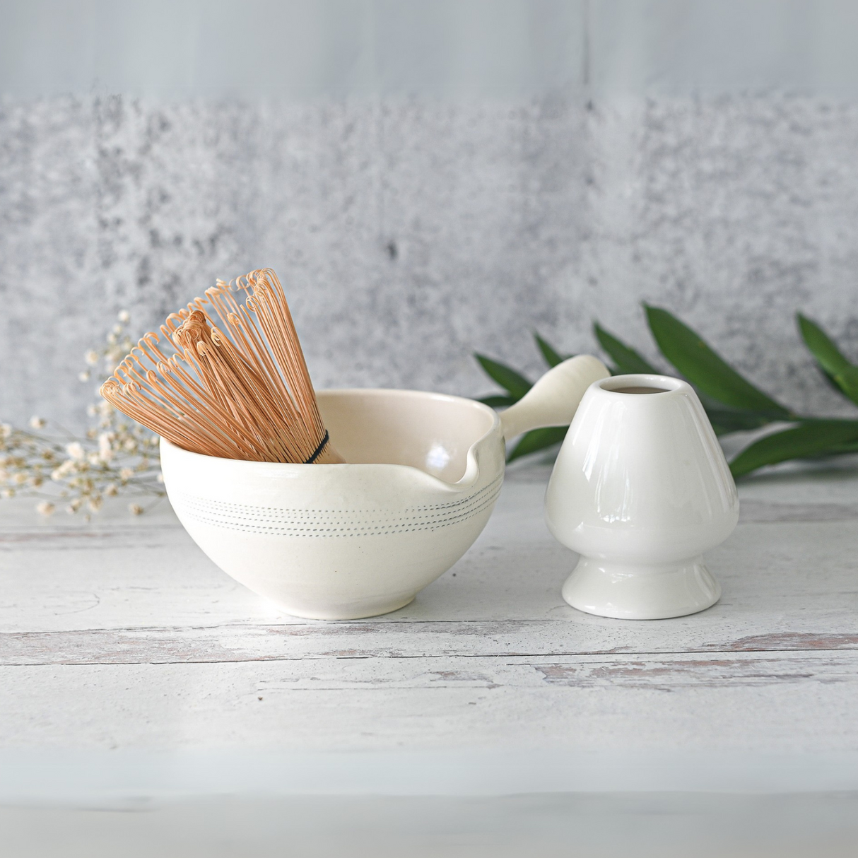 The White Striped Ceramic Matcha Bowl with Spout and Optional Set includes a bamboo whisk and holder, ideal for an authentic matcha tea ceremony on a textured surface.