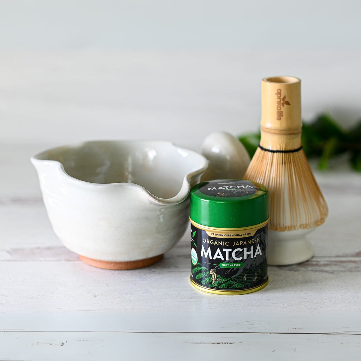Explore the art of matcha with the White Wavy Ceramic Matcha Bowl with Spout, complete with a bamboo whisk and a tin of organic Japanese matcha, beautifully showcased on a pristine white wooden surface.