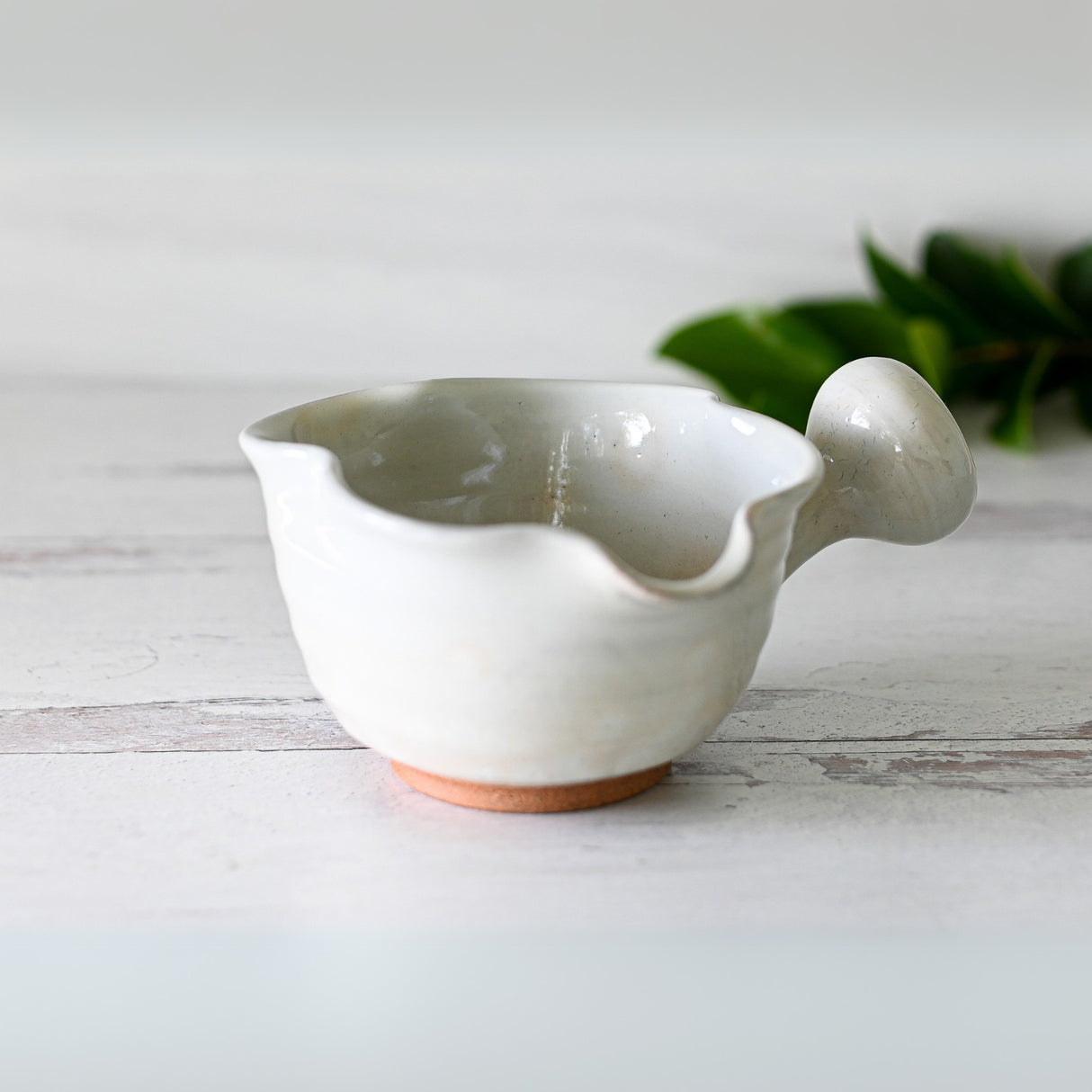 A White Wavy Ceramic Matcha Bowl with Spout, from the Organic Japanese Matcha Set, rests elegantly on a wooden surface, reminiscent of Minoyaki pottery. Blurred green leaves provide a calming background.