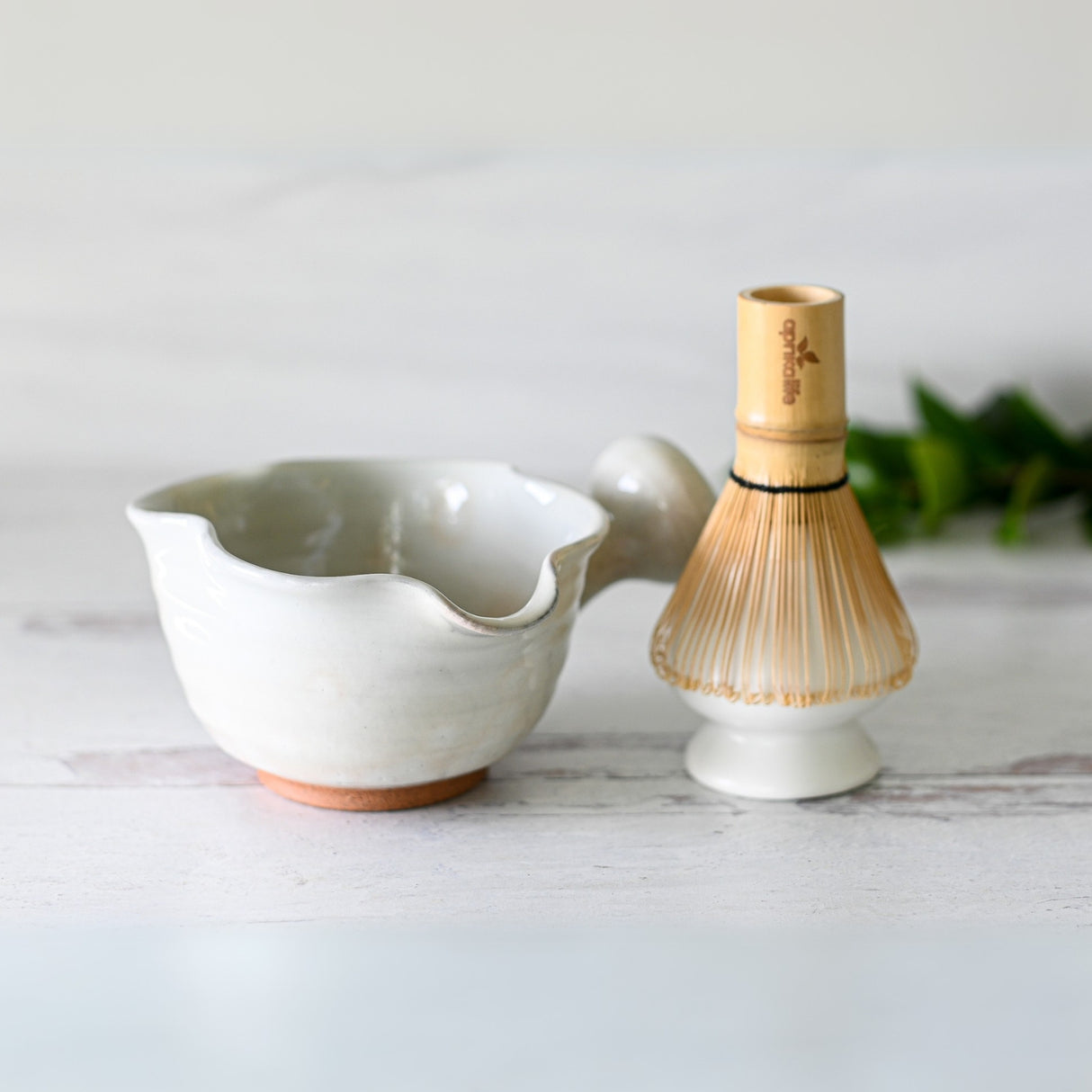 A White Wavy Ceramic Matcha Bowl with Spout sits next to a Bamboo Matcha Whisk on a light surface.