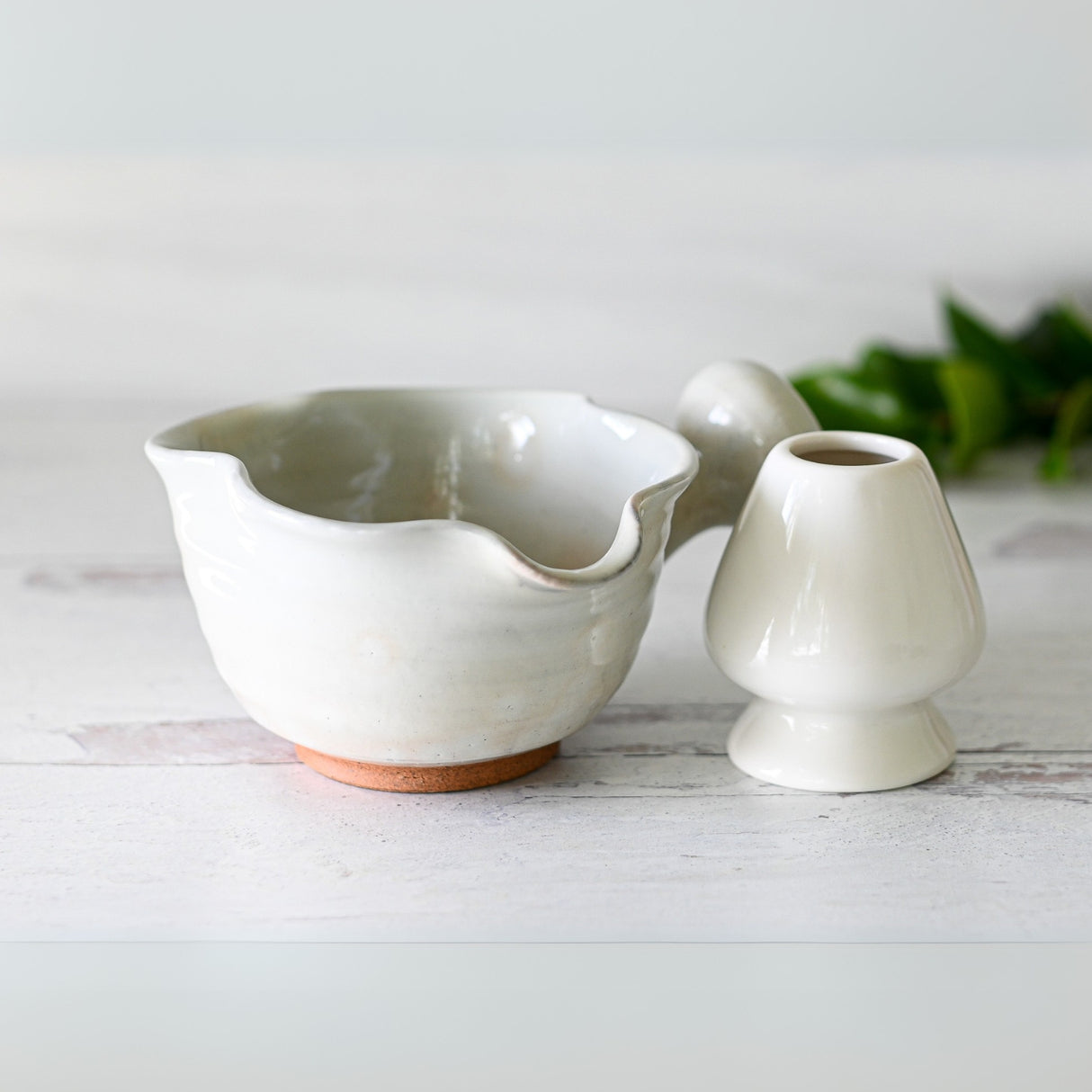 A White Wavy Ceramic Matcha Bowl with Spout sits on a light wooden surface, evoking the charm of traditional Japanese matcha bowls, while a small plant adds serenity to the scene.