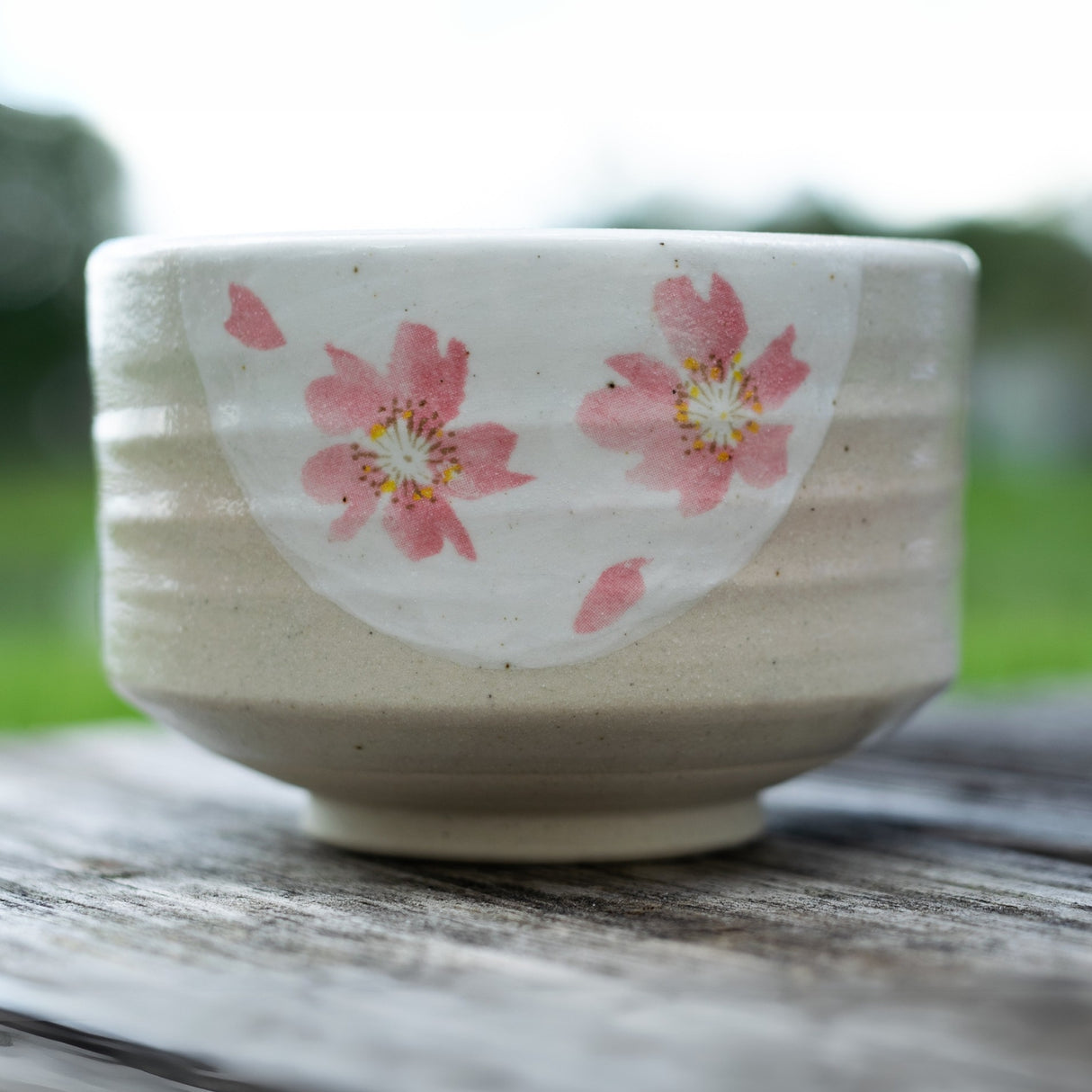 The Beige with Pink Flowers Matcha Bowl, Bamboo Matcha Whisk and Whisk Holder Set is displayed on a wooden surface outdoors, its elegant floral design inspired by traditional artistry.