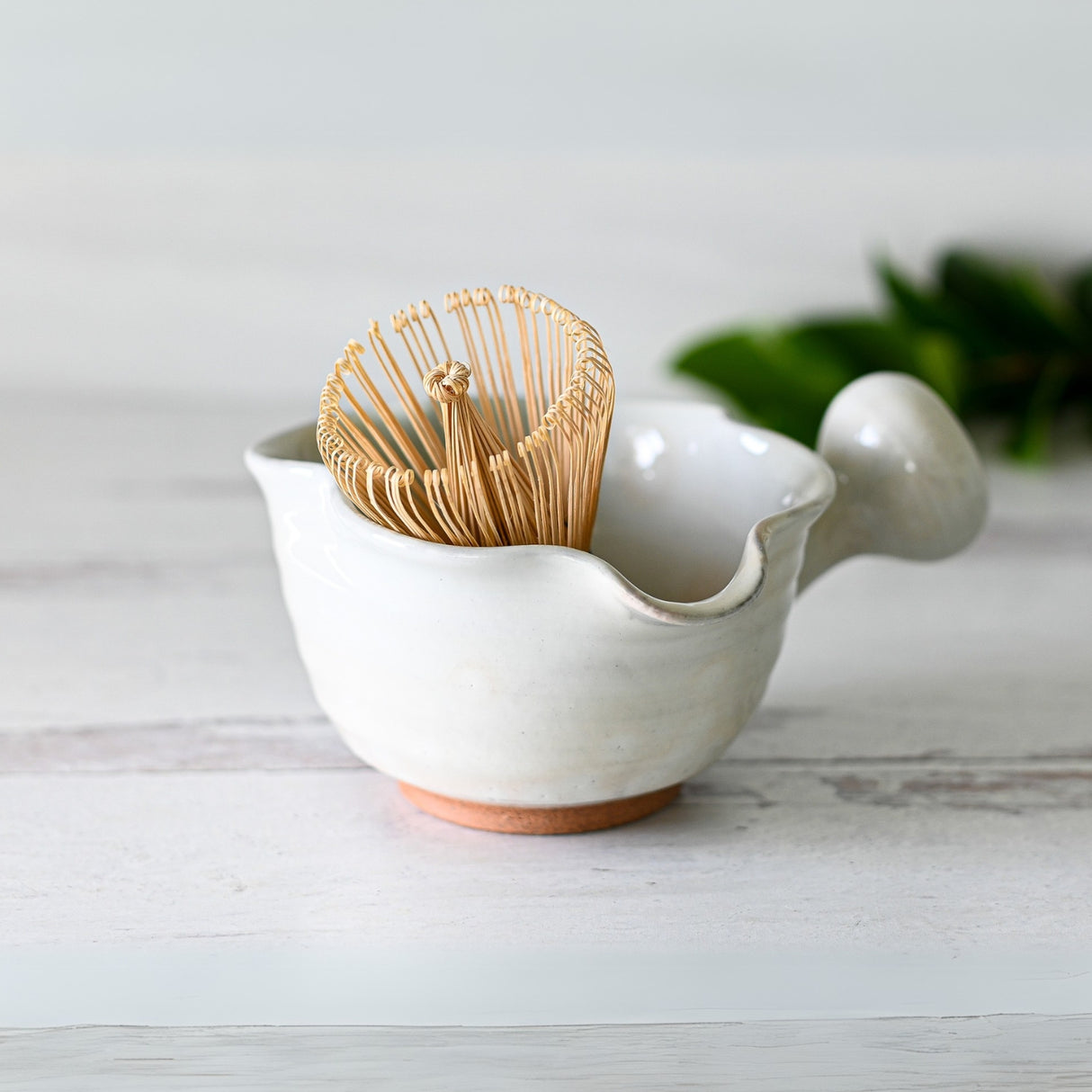 On a light wooden surface sits the White Wavy Ceramic Matcha Bowl with Spout, perfectly complemented by a bamboo whisk, epitomizing a harmonious matcha tea set.