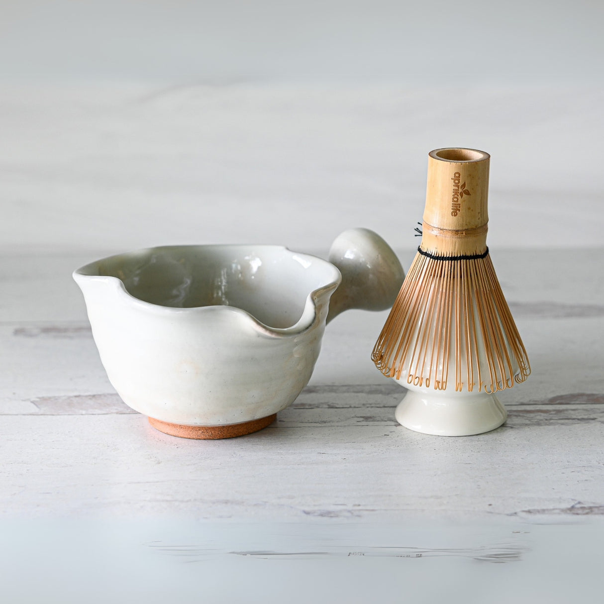 The White Wavy Ceramic Matcha Bowl with Spout and a bamboo whisk create an elegant matcha tea set on a light wooden surface.