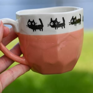 A hand holds a Handmade Pink Ceramic Tea or Coffee Mug from Japan (8oz/250ml), featuring a playful black cats print.