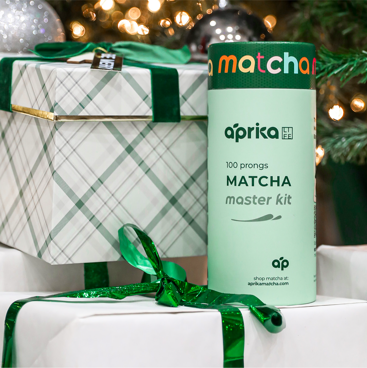 A Matcha Bamboo Whisk & Holder Set, with a bamboo scoop and whisk, sits alongside wrapped gifts under the Christmas tree, adorned with green and plaid accents.