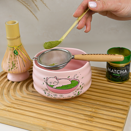 A hand sifts matcha powder into a Cat Print Bowl from the Pink Unfurl Matcha Gift Set, resting on a wooden tray.