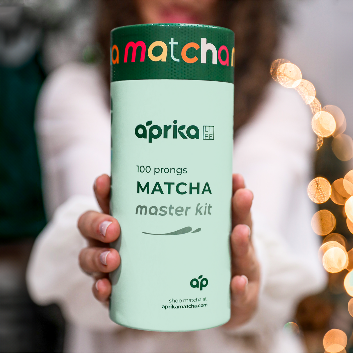 Someone holds an "aprika Matcha Bamboo Whisk & Holder Set" against a festive backdrop, with a bamboo scoop beside it.