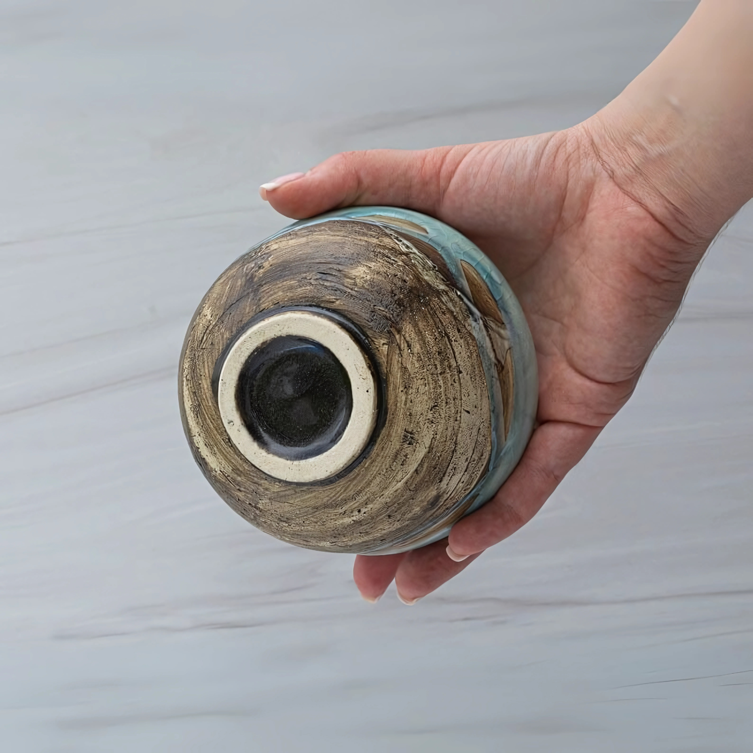A hand holds a Japanese Matcha Tea Bowl Blue & Brown Glaze upside down, displaying its textured, unglazed base against a light background.