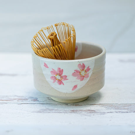 This beautifully crafted beige matcha bowl with pink flowers perfectly complements the bamboo whisk within the Organic Japanese Matcha Set, creating an ideal accessory for preparing matcha on a white wooden surface.