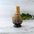 A Black Matcha Whisk Holder and Bamboo Whisk Set sits on a wooden surface, with green leaves in the background, capturing a serene Japanese aesthetic.