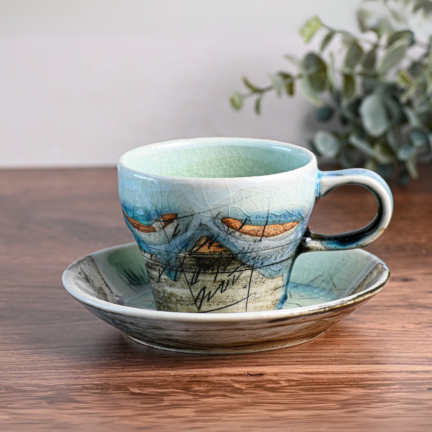 The Blue Ceramic Tea Cup with Saucer, a traditional Japanese tea cup of premium quality ceramic mugs, sits on a wooden table with a nature design and eucalyptus leaves in the background, showcasing exquisite craftsmanship.