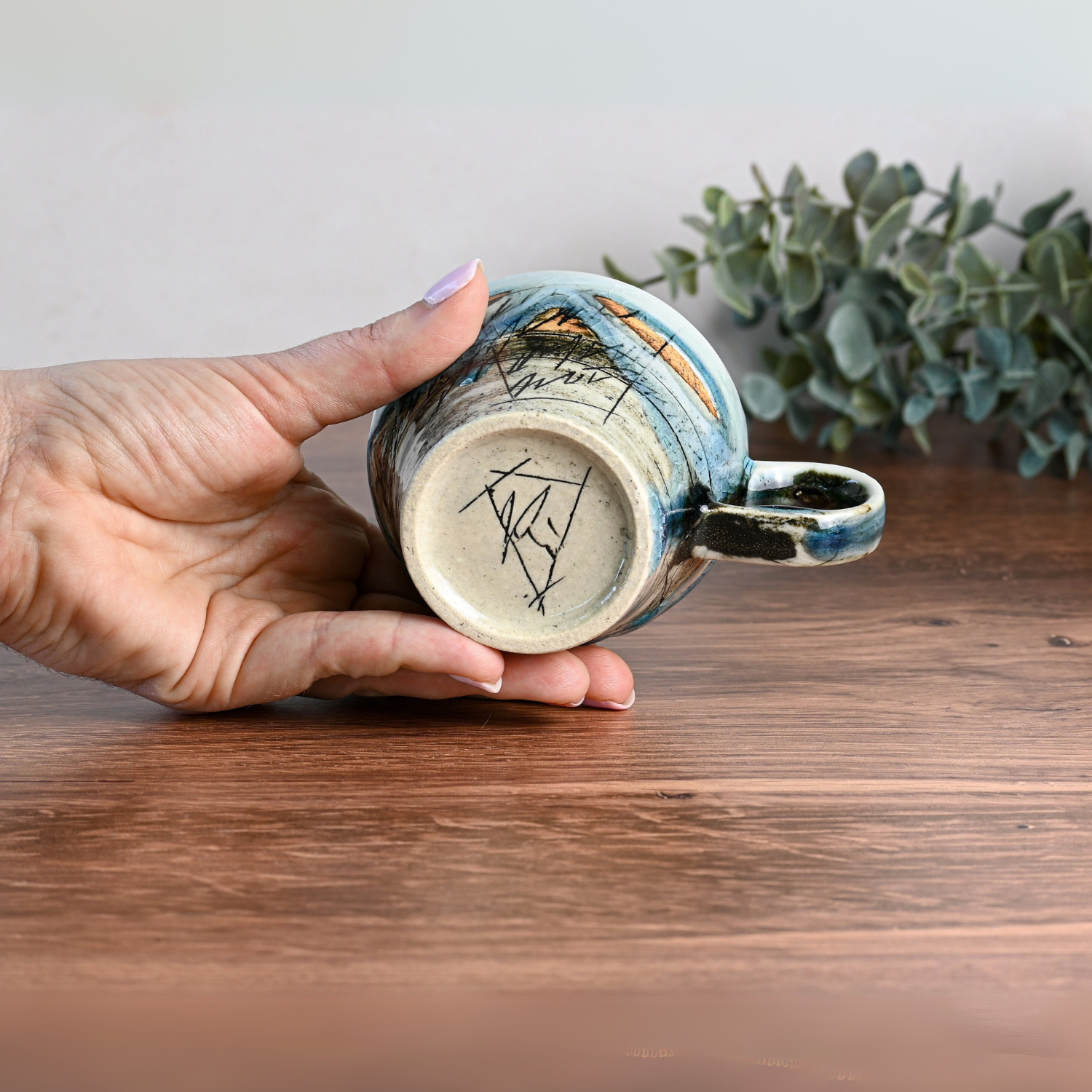 A hand delicately holds a Blue Ceramic Tea Cup with intricate artwork and signatures on the bottom against a wooden surface and lush greenery.
