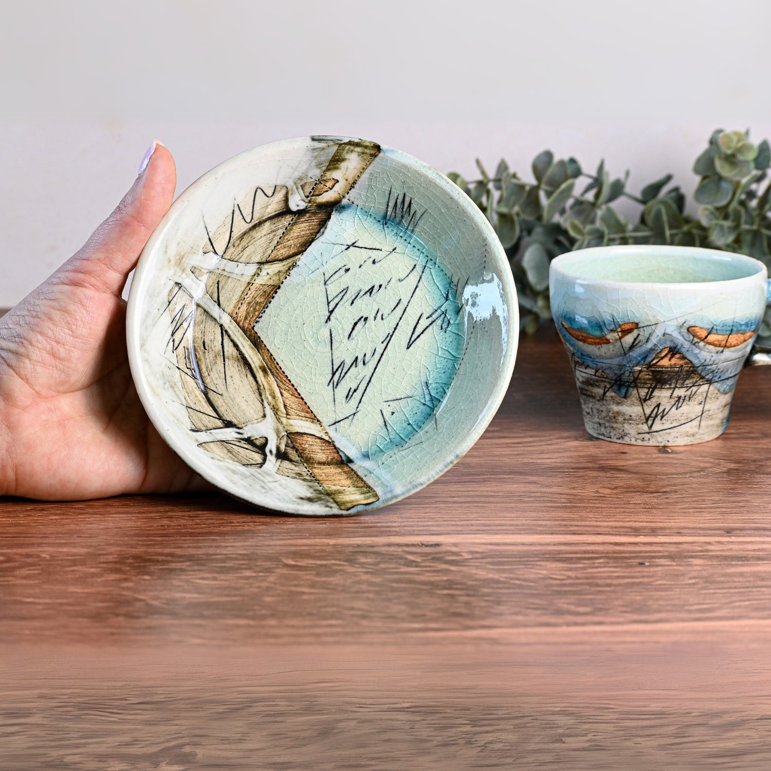 A hand holds a decorative ceramic dish with a Blue Ceramic Tea Cup in the background on a wooden table, showcasing exquisite Japanese craftsmanship.