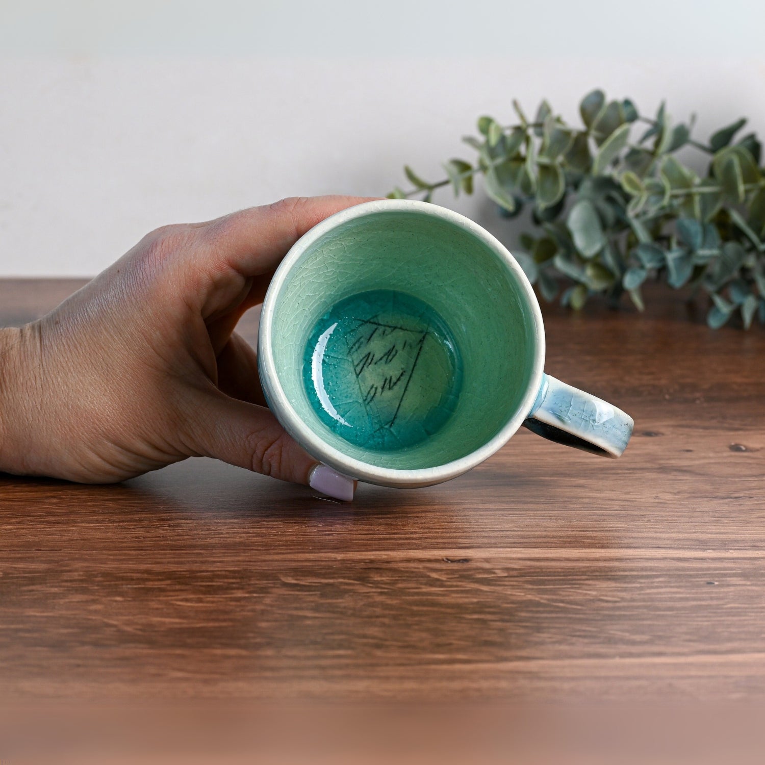 A hand holds a Blue Ceramic Tea Cup with a saucer, showcasing its blue-green interior and accompanied by a handwritten note, all resting on a wooden surface.