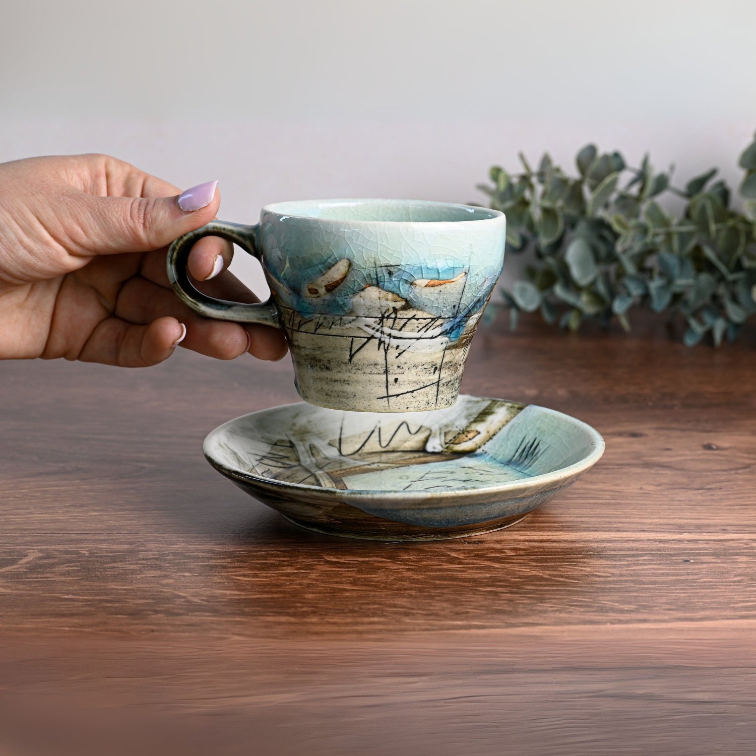 A hand gently cradles a Blue Ceramic Tea Cup with Saucer on a wooden table. Eucalyptus in the background complements the artistry of this Traditional Japanese Tea Cup, showcasing premium craftsmanship.