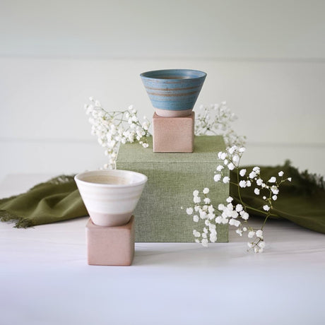 Two blue coffee mugs on pink pedestals, with white flowers and green fabric in the background, showcase exquisite ceramic artistry.