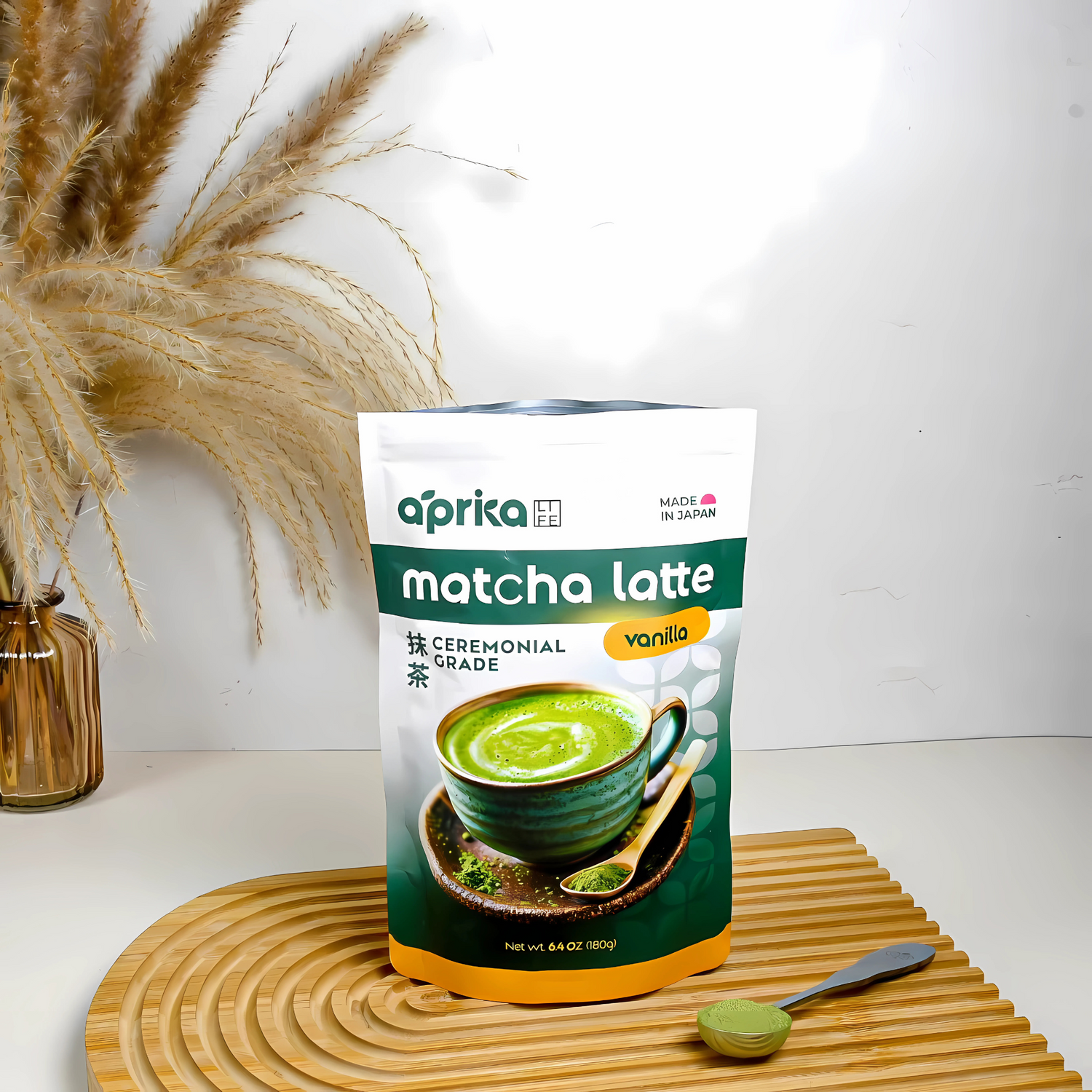 A bag of Ceremonial Grade Matcha Latte Powder Mix with Natural Vanilla Flavor sits on a wooden mat, accompanied by a matcha whisk and pampas grass in the background.
