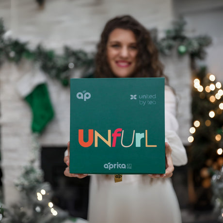A person beams in front of a festive backdrop, holding a green "Unfurl" box—a perfect addition to the Ceremonial Matcha Starter Kit - Black.