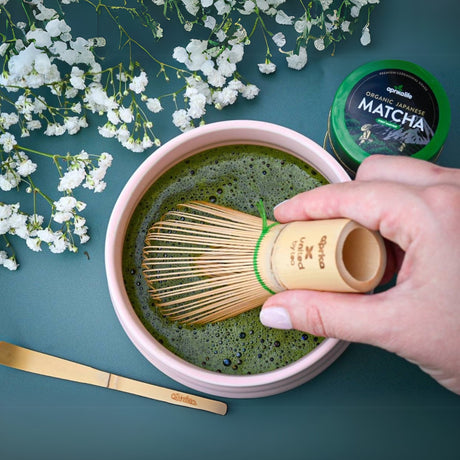 Using the Ceremonial Matcha Starter Kit - Ivory White, whisk matcha in a pink ceramic bowl surrounded by delicate white flowers with a bamboo whisk.