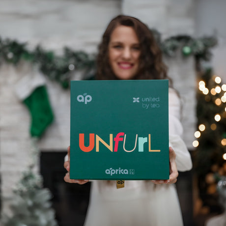 A smiling person holds a green "UNfuRL" box against a festive backdrop. Inside is the "Ceremonial Matcha Starter Kit - Ivory White," featuring a Bamboo Whisk and Ceramic Matcha Bowl, ideal for adding warmth to holiday celebrations.