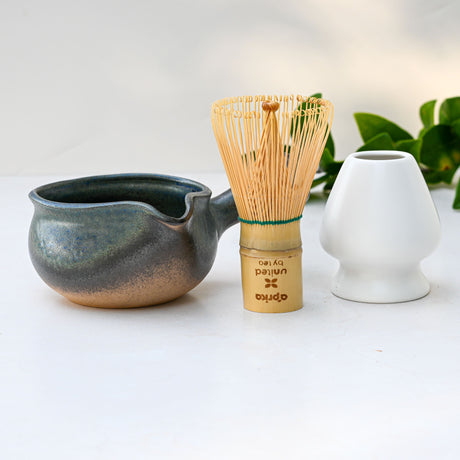 Blue Spouted Ceramic Matcha Bowl, bamboo matcha whisk, and white vase on a light surface with green leaves in the background—each item beautifully handcrafted in Japan.