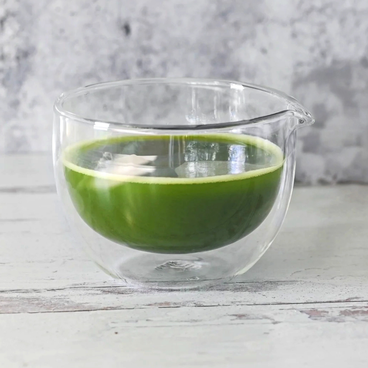 A Double Sided (Double Wall) Glass Matcha Bowl with Spout, filled with green juice, sits on a light wooden surface against a gray background.