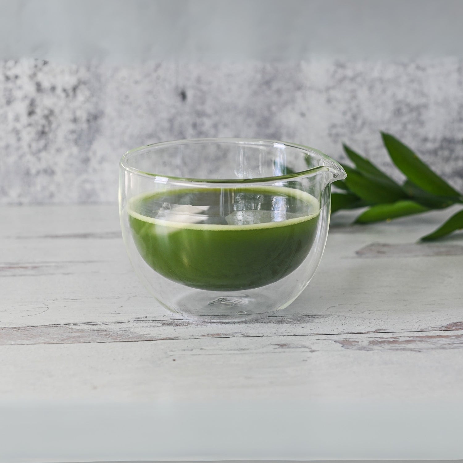A vibrant green matcha latte is elegantly cradled in a Double Sided Glass Matcha Bowl with Spout on a white wooden surface, accompanied by freshly placed green tea leaves.