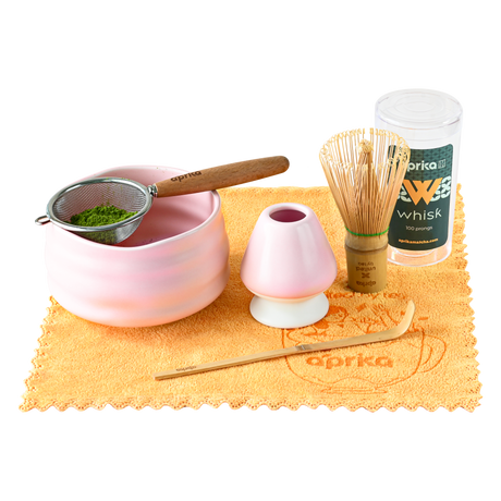 The Ceremonial Matcha Starter Kit - Pink includes a bamboo whisk, strainer, ceramic matcha bowl, scoop, and a pink holder on an orange cloth.