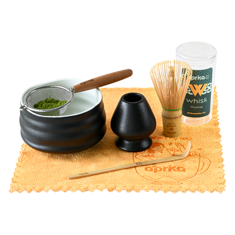 The Ceremonial Matcha Starter Kit - Black includes a black bowl, bamboo whisk, whisk holder, tea strainer, scoop, and container arranged elegantly on a yellow cloth.
