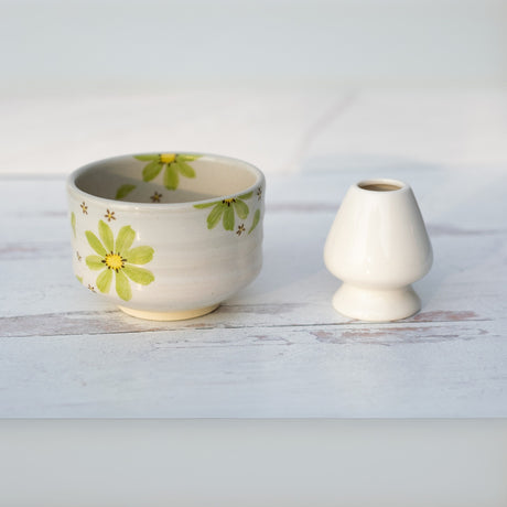 A Green Flowers Ceramic Matcha Bowl from the Matcha Set, featuring a floral design, sits beside a plain white vase on a light surface, inspired by traditional Japanese pottery.