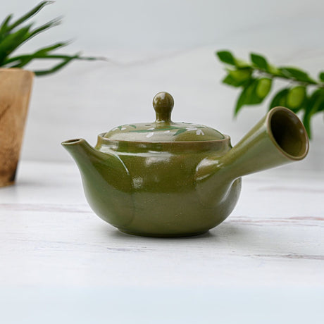 The Green Kuysu with Flowers, a 14 fl oz Japanese handmade teapot featuring a premium tea strainer, graces the white surface adorned with green plants in the background.