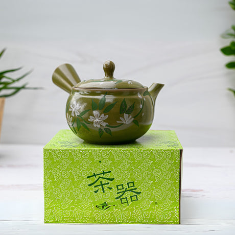 The Green Kuysu teapot, handmade with a floral design and premium tea strainer (14 fl oz/420ml), sits in a patterned lime green box, its artistry reflecting Japanese tradition against an elegant plant backdrop.