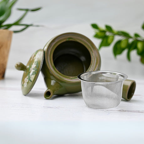 The Green Kuysu with Flowers, a Japanese handmade teapot with a premium tea strainer, lies on its side against a white surface with a blurred plant in the background, highlighting its nod to Tokoname pottery.