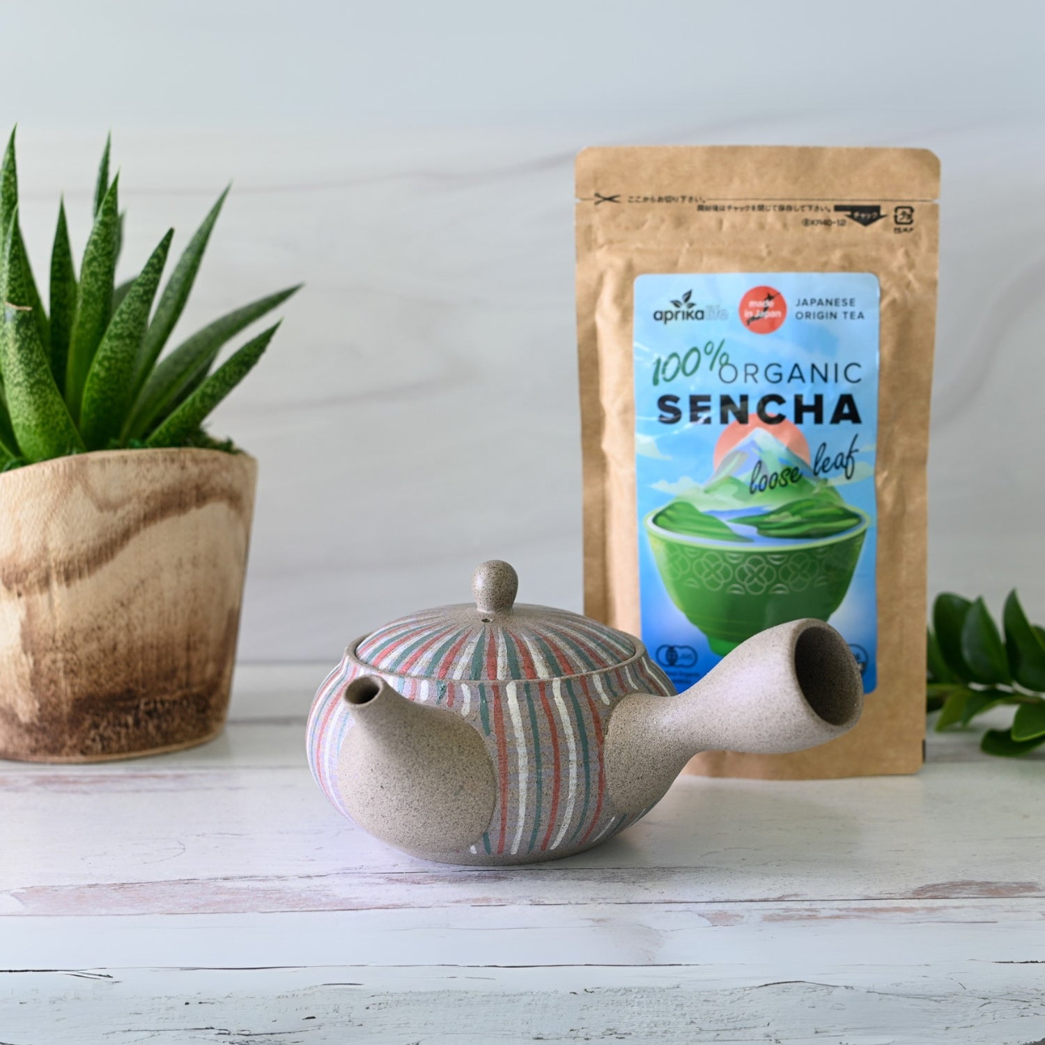 A Handcrafted Japanese Kyusu Teapot with Filters (310ml) and a bag of organic sencha tea rest elegantly on a wooden surface, accompanied by a potted plant, epitomizing the art of Japanese ceramics.
