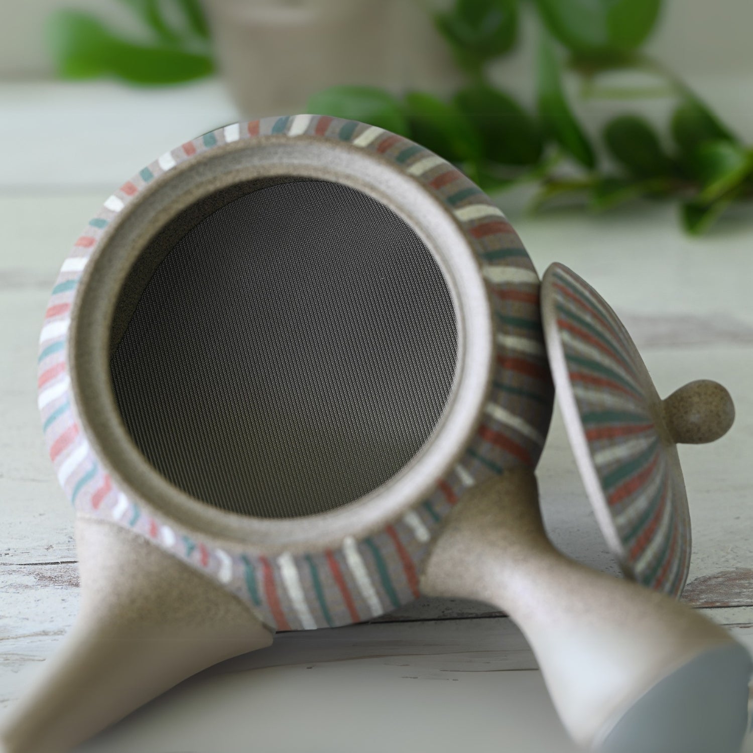The Handcrafted Japanese Kyusu Teapot with Filters (310ml) sits on a light wooden surface, its lid removed and green leaves as a serene backdrop. With colorful stripes, it embodies the essence of traditional Tokoname pottery.