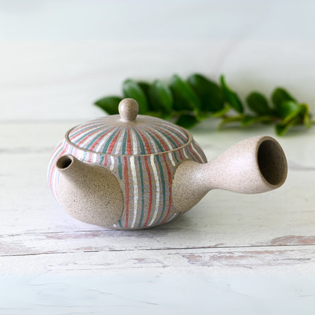 The Handcrafted Japanese Kyusu Teapot with Filters (310ml), adorned in vibrant stripes, gracefully sits on a white wooden surface, framed by lush green leaves.