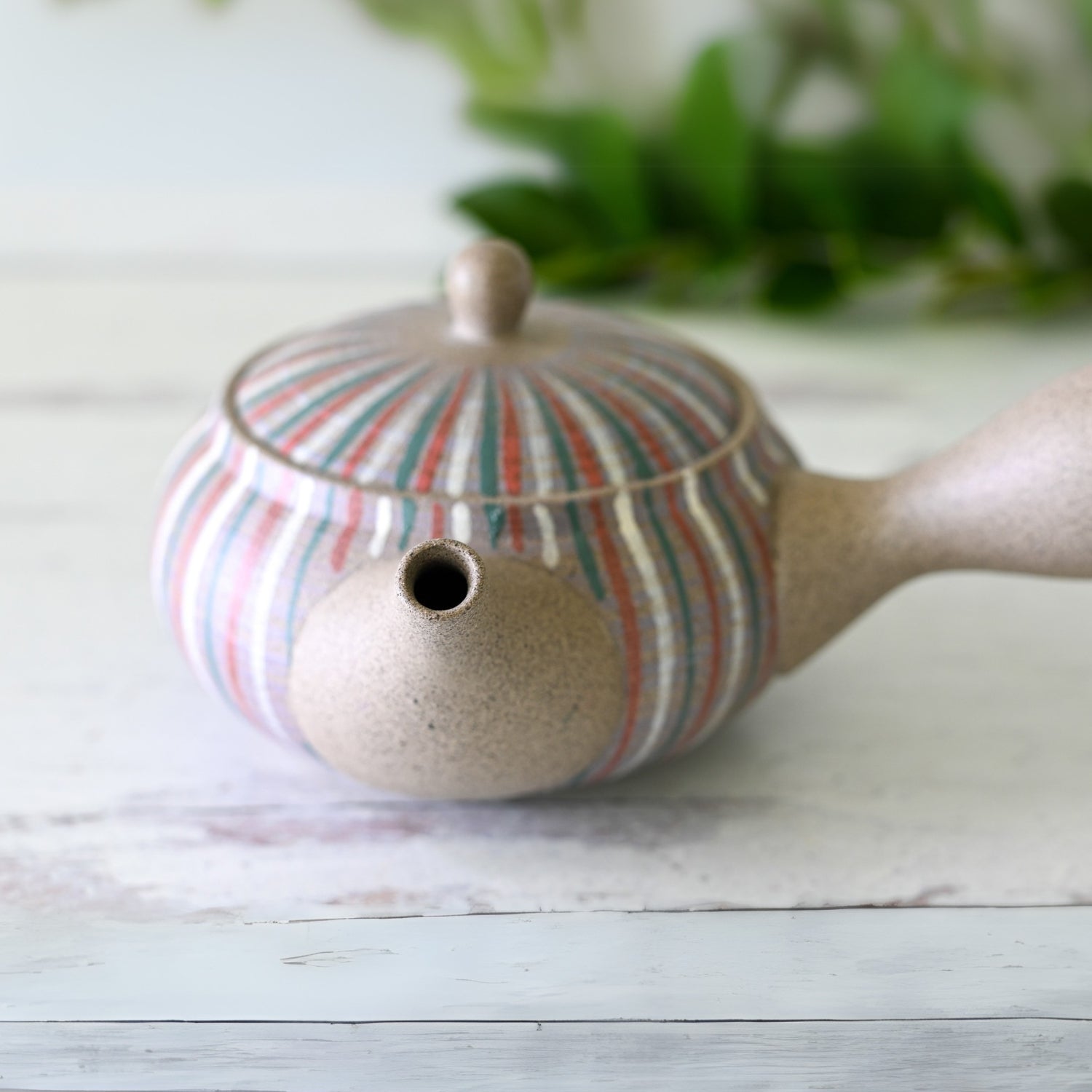 A Handcrafted Japanese Kyusu Teapot with Filters (310ml) featuring a spout and a striped design sits on a light wooden surface, lush greenery in the background.
