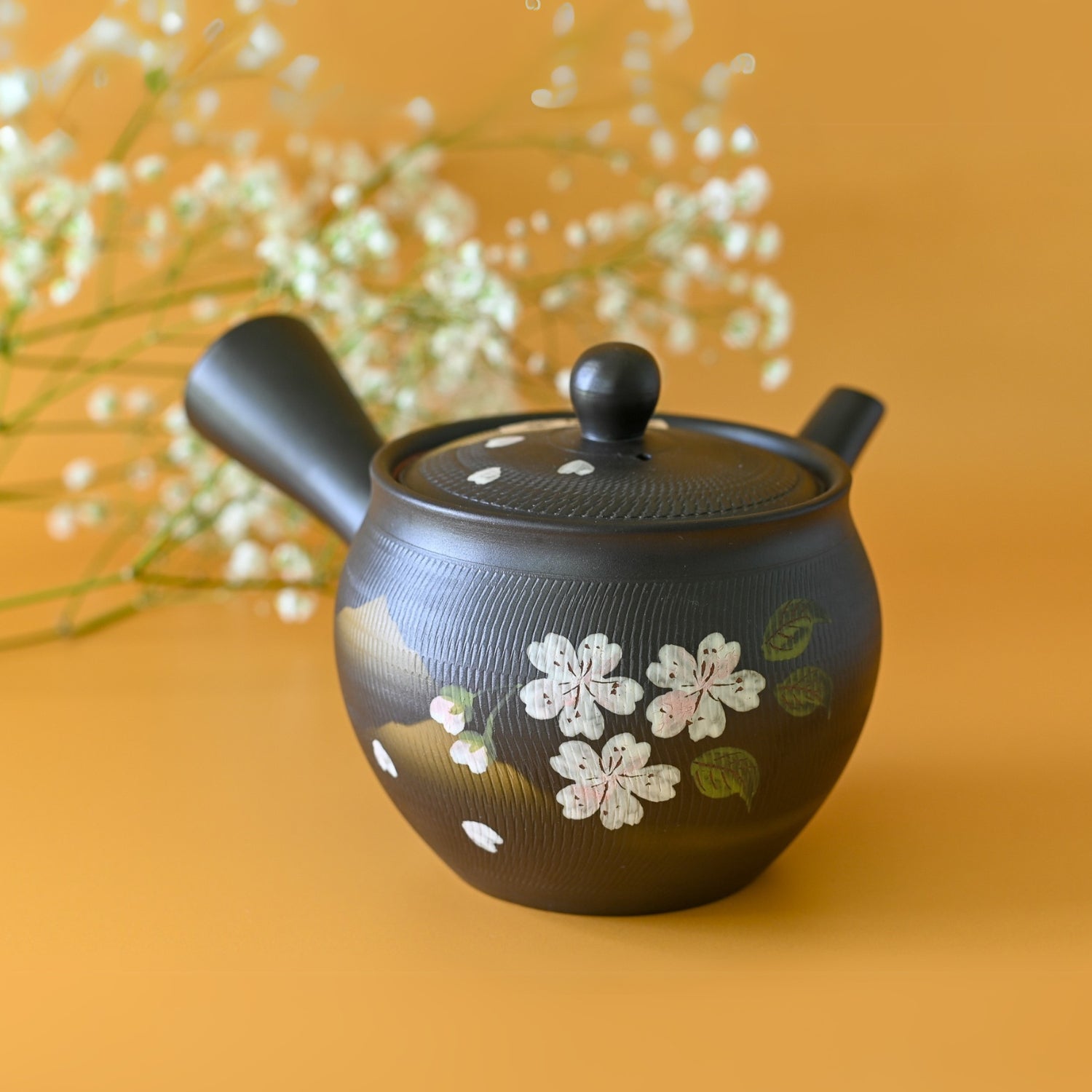 The Handcrafted Japanese Kyusu Teapot with Filters (350 ml) is a black teapot with floral designs on a yellow base, capturing the elegance of Japanese ceramics. It features delicate white flowers and is reminiscent of a Deep Steaming Teapot made from Tokoname clay.