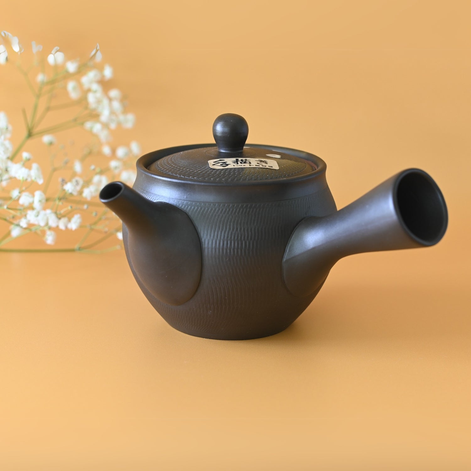 The Handcrafted Japanese Kyusu Teapot with Filters (350 ml) in black Tokoname clay, featuring a side handle, is elegantly placed on a yellow surface beside white flowers.