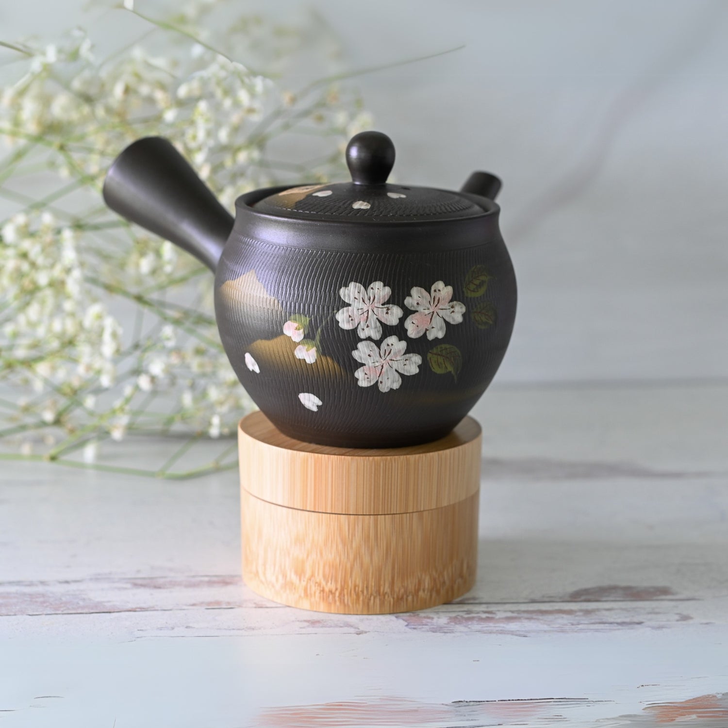 The Handcrafted Japanese Kyusu Teapot with Filters (350 ml) features a black ceramic design adorned with floral patterns. It’s elegantly displayed on a wooden stand, surrounded by white flowers, demonstrating the craft of Tokoname clay artistry.