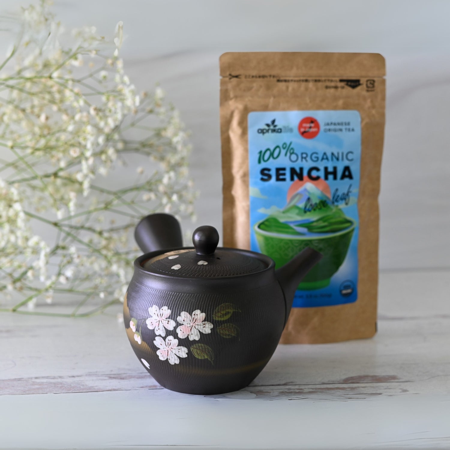 A Handcrafted Japanese Kyusu Teapot with Filters (350 ml), made from Tokoname clay with a floral design, sits on a light surface alongside a 100% organic sencha tea bag and delicate white flowers in the background.