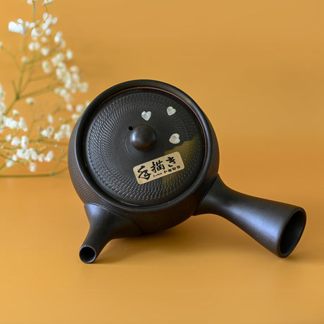 The Handcrafted Japanese Kyusu Teapot with Filters (350 ml), made from Tokoname clay and adorned with small heart shapes, sits on an orange surface. White flowers are in the background.