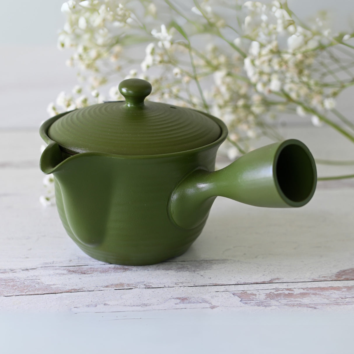 The Handcrafted Japanese Kyusu Teapot with Filters (380ml) sits elegantly on a white wooden surface, accompanied by delicate white flowers in the background. Its design evokes Fujita Tokuta's artistry, blending functionality with timeless beauty.