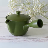 The Handcrafted Japanese Kyusu Teapot with Filters (380ml) sits elegantly on a white wooden surface, accompanied by delicate white flowers in the background. Its design evokes Fujita Tokuta's artistry, blending functionality with timeless beauty.