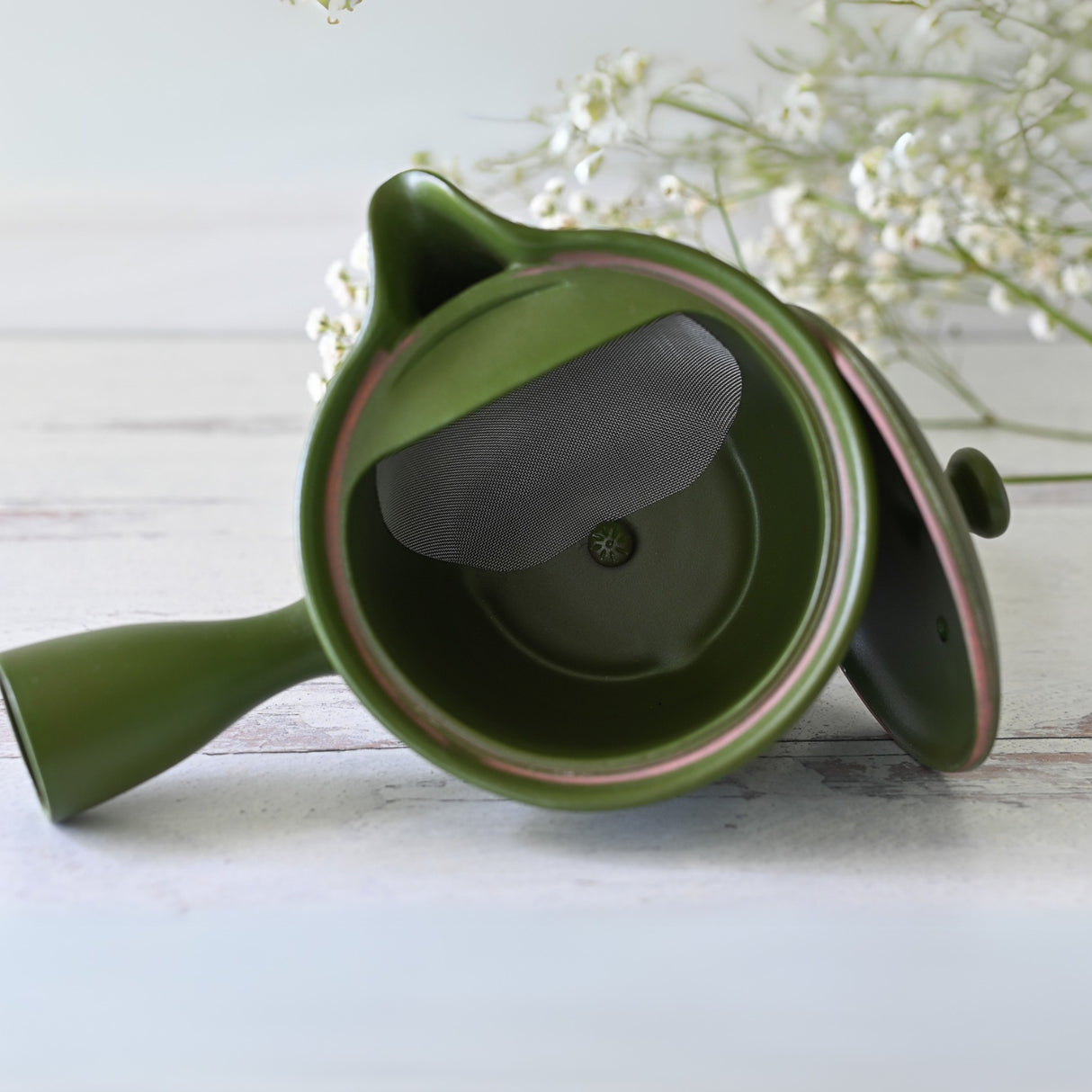 A Handcrafted Japanese Kyusu Teapot with Filters (380ml) in green rests on a white wooden surface, accompanied by small white flowers, capturing the elegance of Japanese ceramics.